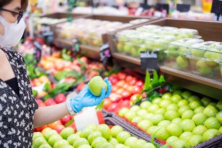 Asian woman wearing nitrile gloves and holding green apple fruit in supermarket or grocery, protect coronavirus inflection. Hygiene, new normal and life after covid-19 pandemicの写真素材