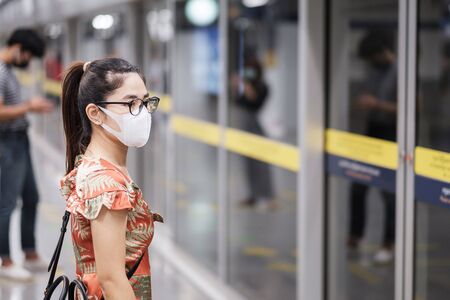 young Asian girl wearing Surgical face mask protect coronavirus inflection, Happy tourist woman wating public train. social distancing, new normal and life after covid-19 pandemicの写真素材