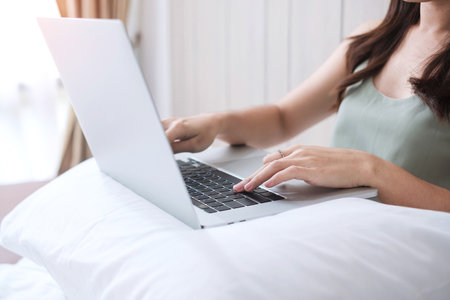 Happy casual woman sitting and using computer laptop on white pillow in bedroom. Young adult female Working from home or online shopping in morning. Freelancer, lifestyle and technology conceptsの写真素材
