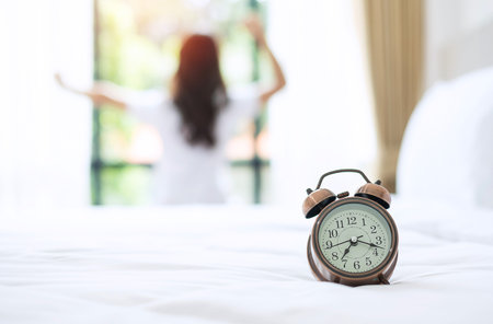 Closeup alarm clock with background of happy woman stretching in bed after waking up, young adult female rising arms and looking to window in the morning. fresh relax and have a nice day conceptsの写真素材