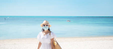 Happy tourist woman wearing surgical face mask  enjoy beautiful sea and white sand at tropical beach. protection Coronavirus disease (Covid-19) infection. New Normal, travel, vacation and holidayの写真素材