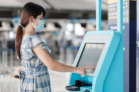 woman wearing medical face mask and using the self check-in machine at the airport terminal getting...