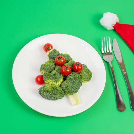 Christmas tree of organic vegetables on white plate; Broccolis and tomatoes with fork and knife on green background. Merry Xmas and happy New Year party, New You, Healthy food and vegetarian conceptの写真素材