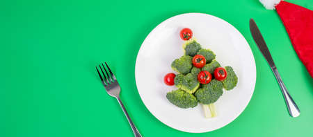 Christmas tree of organic vegetables on white plate; Broccolis and tomatoes with fork and knife on green background. Merry Xmas and happy New Year party, New You, Healthy food and vegetarian conceptの写真素材