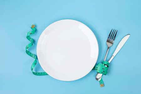 Top view Green Measuring tape wrapped around fork and knife with white ceramic dish on blue...