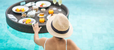 Happy woman in white swimsuit having Floating Breakfast tray in luxury pool hotel, young female with...