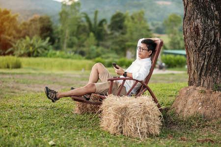 Young blogger man using smartphone during relaxing in garden, asian freelancer contracting business online. Digital nomad, freedom and technology conceptの写真素材