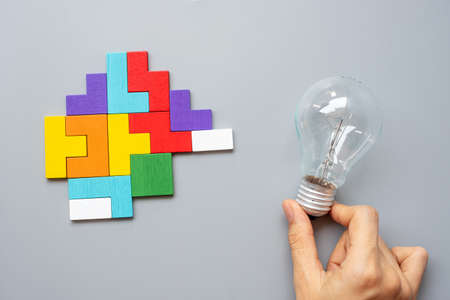 Hand holding lightbulb with colorful wood puzzle piece on gray background.の写真素材