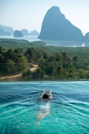 Happy woman in white swimsuit swimming in luxury pool hotel against Phang Nga bay background,...
