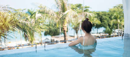 Happy woman in white swimsuit swimming in luxury pool hotel, young female with hat enjoy in tropical...