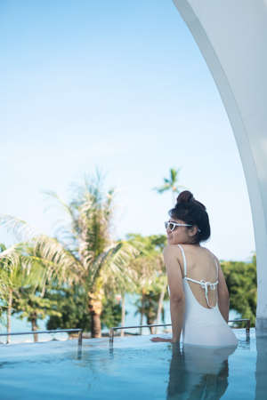 Happy woman in white swimsuit swimming in luxury pool hotel, young female with hat enjoy in tropical...