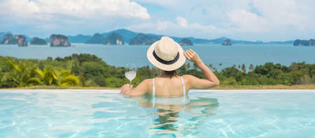 Happy woman in white swimsuit drinking wine in luxury swimming pool hotel against beautiful view, young female with hat enjoy in tropical resort. Relaxing, summer travel, holiday and vacation conceptの写真素材