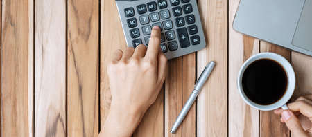 Top view Business woman hand holding coffee cup and calculating with calculator, pen and computer laptop on wood office desk table background. workspace or home office with copy space for text conceptの写真素材