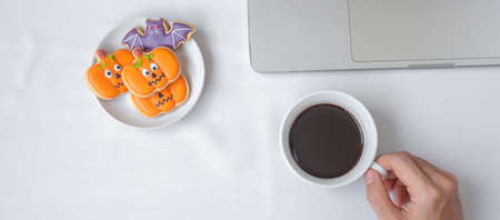 Hand holding coffee cup and eating Halloween Cookies during using computer laptop. Happy Halloween, online shopping, Hello October, fall autumn, Festive, party and holiday conceptの写真素材