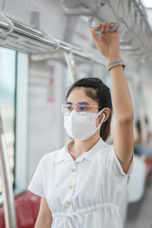 woman with Medical face mask, Hand holding handle and listening music by earphones in train. public transportation and safety under covid-19 pandemicの写真素材
