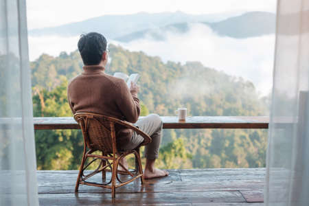 young man reading book near window and looking mountain view at countryside homestay in the morning sunrise. SoloTravel, journey, trip and relaxing conceptの写真素材