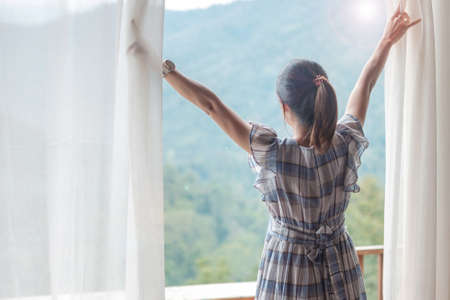 happy woman stretching near window, young adult female rising arms and looking mountain view in the morning. relaxing and lifestyle conceptsの写真素材