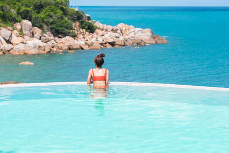 Happy woman in orange swimsuit swimming in infinity pool at luxury hotel against ocean front. young female enjoy in tropical resort. Relaxing, summer,  travel, holiday, vacation and weekend conceptの写真素材