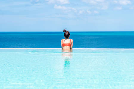 Happy woman in orange swimsuit swimming in infinity pool at luxury hotel against ocean front. young female enjoy in tropical resort. Relaxing, summer,  travel, holiday, vacation and weekend conceptの写真素材