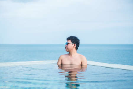 Happy man in infinity pool at luxury hotel against ocean front. young male enjoy in tropical resort. Relaxing, summer,  travel, holiday, vacation and weekend conceptの写真素材