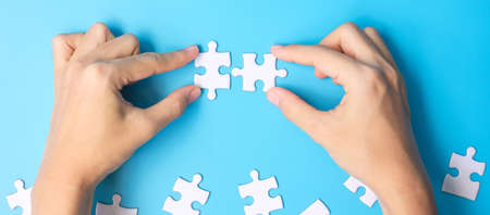 hands connecting couple white puzzle jigsaw pieces on blue background. Concept of solutions, mission, success, goals, cooperation, partnership, strategy and puzzle dayの写真素材