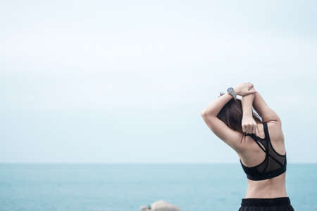 Young fitness female in sportswear stretching body against ocean view, healthy woman exercise in morning. Workout, wellness and work life balance conceptsの写真素材
