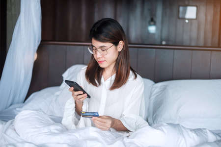 woman using mobile smart phone and credit card for online shopping while making order on bed in morning at home. technology, ecommerce, digital banking online payment and apartment living conceptの写真素材
