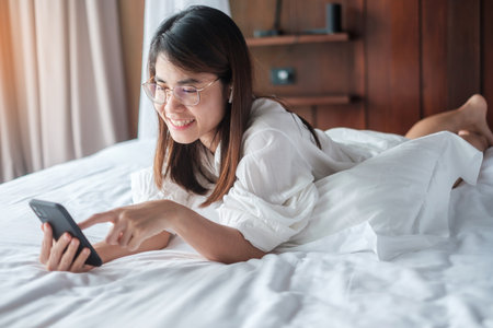 woman using smartphone for social media, young female watching movies by mobile phone on bed at home. technology, network, online shopping, e commerce, lifestyle and digital communication conceptの写真素材