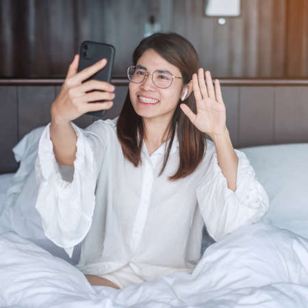 Happy woman using smart phone for video call, female meeting  online by mobile phone on bed. technology, network, work from home, lifestyle and digital communication conceptの写真素材