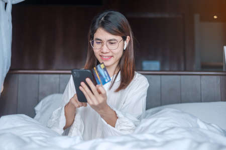 woman using mobile smart phone and credit card for online shopping while making order on bed in morning at home. technology, ecommerce, digital banking online payment and apartment living conceptの写真素材