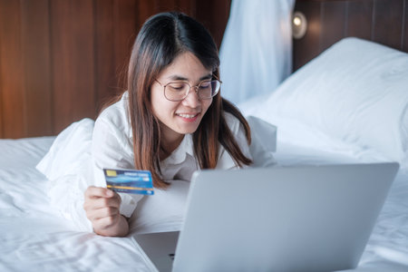 woman using computer laptop and credit card for online shopping while making order on bed in morning at home. technology, ecommerce, digital banking online payment and apartment living conceptの写真素材
