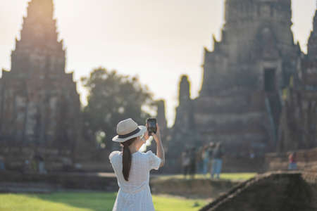 Happy tourist woman in white dress taking Photo by mobile smartphone, during visiting in Wat Chaiwatthanaram temple in Ayutthaya Historical Park, summer, solo, Asia and Thailand travel conceptの写真素材