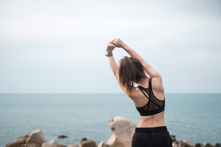 Young fitness female in sportswear stretching body against ocean view, healthy woman exercise in morning. Workout, wellness and work life balance conceptsの写真素材
