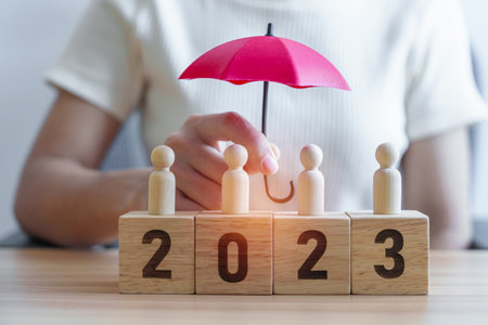 hand holding umbrella cover wooden man employees with 2023 Year block. Insurance, protect, People,...