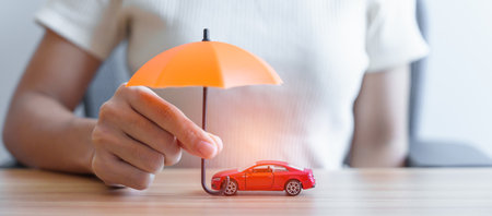 Businesswoman hand holding orange umbrella and cover red car toy on table. Car insurance, warranty, repair, Financial, banking and money conceptの写真素材