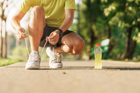 Young athlete man tying running shoes with Energy Drink water, male runner ready for jogging outside, asian Fitness walking and exercise in the park morning. wellness, wellbeing and sport conceptsの写真素材