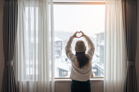 young woman in sweater looking through the window in winter season, happy female rising arms and stretching after waking at apartment or home in the morning. Lifestyle and Relaxing conceptsの写真素材