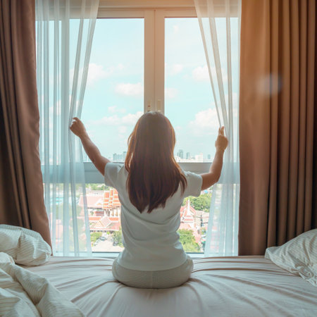 happy woman stretching on bed after wake up, young adult female rising arms and looking to window in...