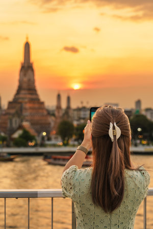 tourist woman enjoys view to Wat Arun Temple in sunset, Traveler take photo to Temple of Dawn by smartphone from rooftop bar. Landmark and Travel destination in Bangkok, Thailand and Southeast Asiaの写真素材