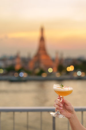 tourist hand shows cocktail glass during enjoy view to Wat Arun Temple in sunset, Traveler visits Temple of Dawn near from rooftop bar. Travel destination in Bangkok, Thailand and Southeast Asiaの写真素材