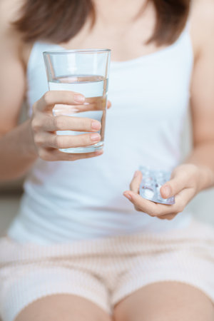 woman hand holding medicine painkiller pill and water glass on the sofa at home, taking for headaches, stomach ache, Diarrhea Pain from food poisoning, Endometriosis, Hysterectomy and Menstrualの写真素材