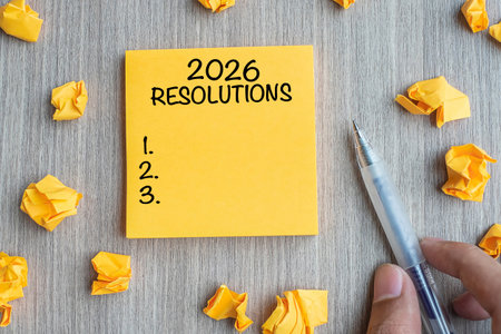 2026 RESOLUTIONS word on yellow note with Businessman holding pen and crumbled paper on wooden table...