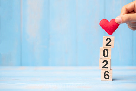 Business man hand holding red heart shape over 2026 wooden cubes on blue table background with copy...