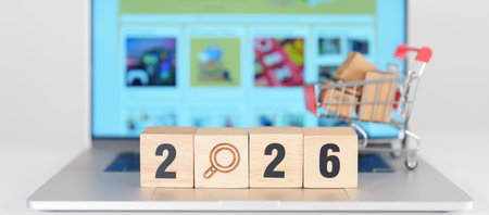 Happy New Year 2026 block with magnifying glasss and shopping trolley cart on table. E commerce,...