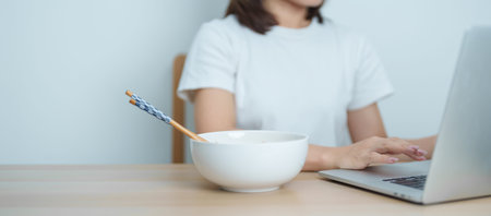 Happy young Asian woman working late at night on laptop while enjoying a bowl of hot instant ramen...