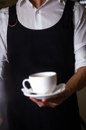 barista in a white shirt and black apron holds a cup of coffeeの写真素材