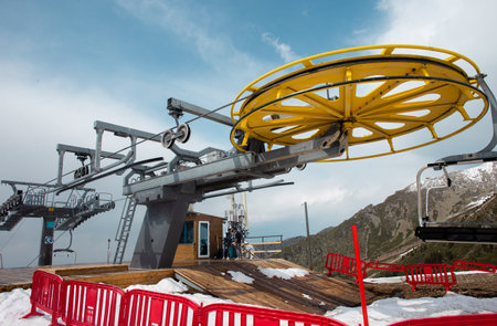 A yellow and gray ski lift wheel and tower with a blue sky and snow-capped mountains in the background. The ski lift is surrounded by red and white striped safety barriers.の写真素材