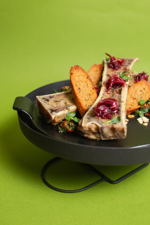 Roasted bone marrow with buttery richness on crusty bread, garnished with fresh herbs, served on a black plate against a green backdrop.の写真素材