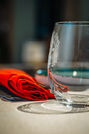A crystal wine glass with a delicate stem, set on a polished wooden table beside a rich red napkin,...