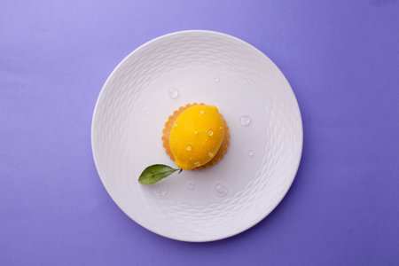 Overhead shot of a hyper-realistic lemon-shaped dessert: textured surface, dew-like droplets, and green leaf on a ridged white plate against purple.の写真素材